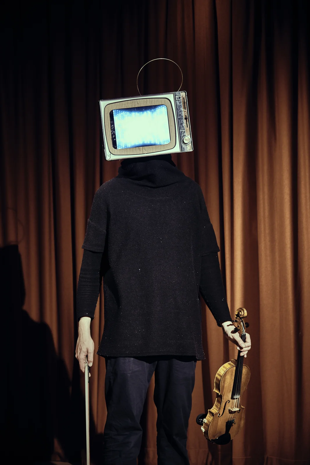 Danny wearing a television head and holding a violin, in front of theatre curtains